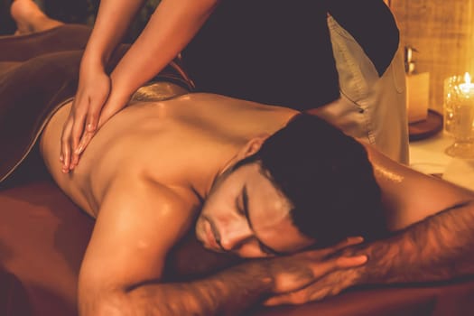 Caucasian man customer enjoying relaxing anti-stress spa massage and pampering with beauty skin recreation leisure in warm candle lighting ambient salon spa at luxury resort or hotel. Quiescent