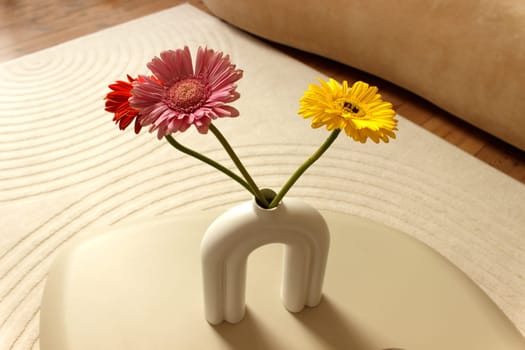 Three colored gerbera flowers in a vase on a table as part of a beige room design. High quality photo