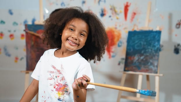 Cute african girl holding painted brush while student drawing canvas behind. Multicultural children attend in art lesson while pretty learner looking at camera. Creative activity concept. Erudition.