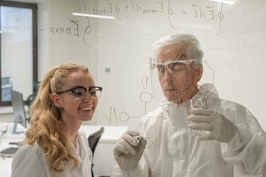 Two chemist colleagues write formulas on glass. Caucasian elderly man and young woman brainstorming