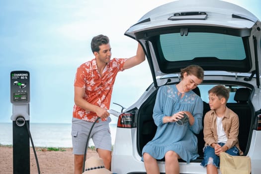 Family vacation trip traveling by the beach with electric car, lovely family sit on the trunk, charging EV car battery with green and sustainable energy. Family travel and eco-friendly car. Perpetual