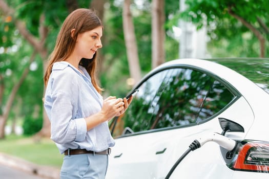 Young woman recharge EV electric vehicle battery from EV charging station and using smartphone online banking to pay for electricity in city park. Eco friendly vehicle travel with EV car. Exalt