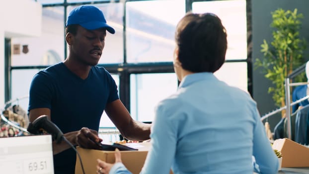 Caucasian worker discussing with courier, after preparing packages for delivery in modern boutique. African american deliveryman showing logistics report on tablet computer. Fashion concept