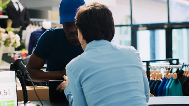 African american courier holding pile with carton boxes, discussing shipping details with store manager in modern boutique. Deliveryman signing shipment report on tablet computer. Fashion concept