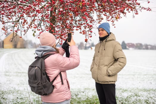 Winter Romance: Girl in Pink Winter Jacket Photographing Boy Against Snow-Covered Red Tree and Field. Embrace the winter magic in this enchanting image, where a girl in a pink winter jacket captures a moment as she photographs her companion against the backdrop of a snow-covered red tree and field. The photograph beautifully conveys the essence of winter romance and the serene beauty of a snowy landscape.