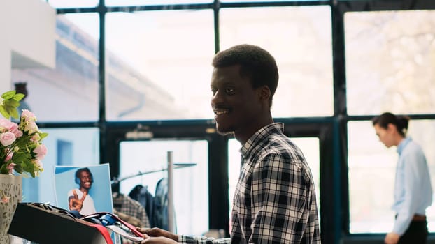 African american stylish man analyzing tie fabric, shopping for fashionable accessories and merchandise in clothing store. Customer looking at shelf with new fashion collection in modern boutique