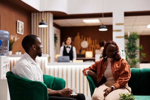 African american guests in lounge area talking and enjoying luxury service at hotel, waiting to fill in registration form for room reservation. Tourists sitting on sofa at front desk.