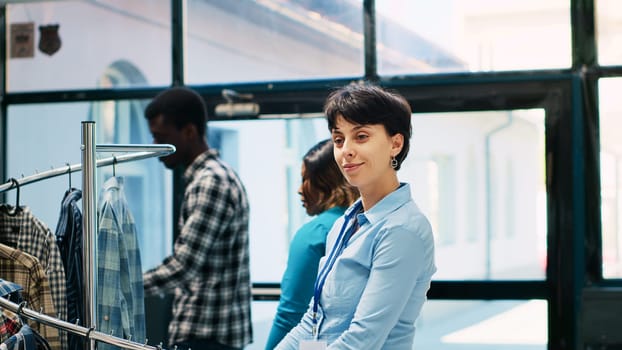 Employee arranging fashionable items in modern boutique, working at store visual. Cheerful worker with short hair looking at new fashion collection, smiling at camera in shopping centre