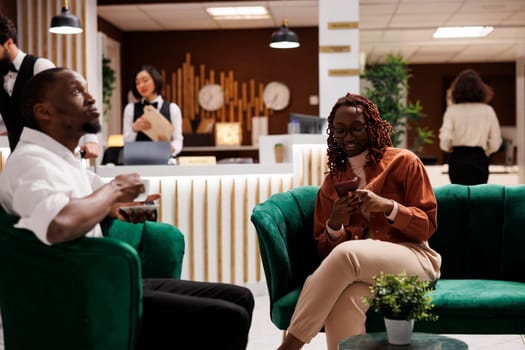 African american woman using smartphone sitting in lounge area at hotel, preparing for room accommodation at luxury resort. Couple waiting to do check in process at reception counter.