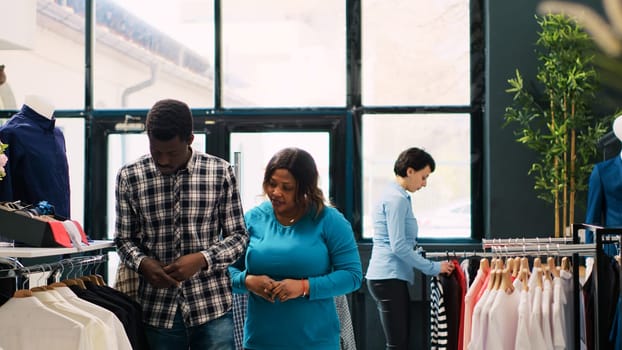 African american couple shopping for trendy clothes, looking at racks full with fashionable merchandise in modern boutique. Stylish customers checking outfit fabric before buying it in clothing store