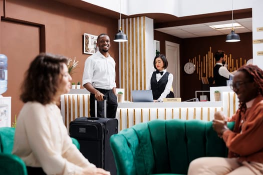 African american guest arriving at hotel, waiting to receive access key and do check in at reception desk. Young adult with luggage preparing to find accommodation, resort lobby.