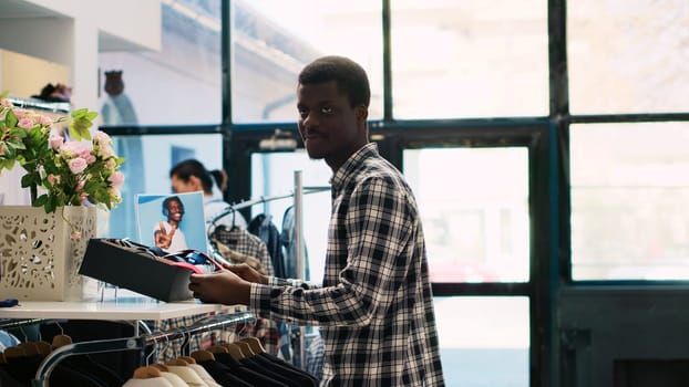 African american customer checking tie material, shopping for stylish accessories and merchandise in clothing store. Man client looking at shelf with new fashion collection in modern boutique