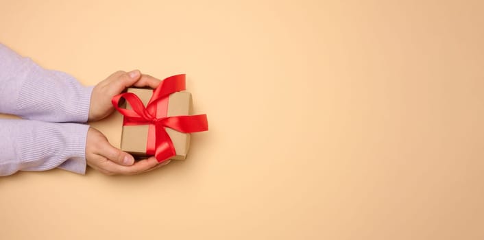 A woman's hand holds a gift box wrapped in a red silk ribbon on a beige background, top view. Copy space