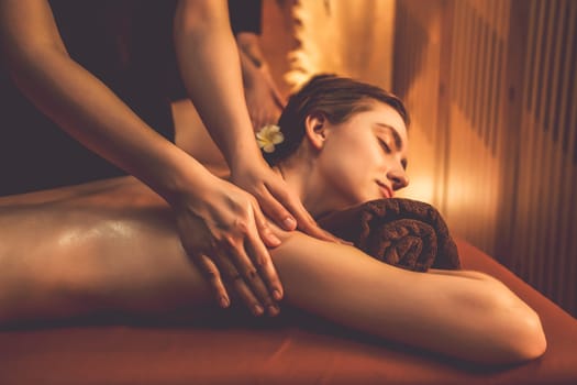 Caucasian couple customer enjoying relaxing anti-stress spa massage and pampering with beauty skin recreation leisure in warm candle lighting ambient salon spa at luxury resort or hotel. Quiescent