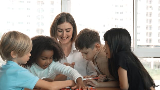 Professional caucasian teacher telling story to diverse student while sitting at table with storybook and colored book. Smart learner listening story while colored picture from instructor. Erudition.