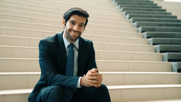 Smiling skilled businessman looking at camera while sitting on stairs. Young professional project manager smile at camera while holding mobile phone at outdoor with blurred background. Exultant.