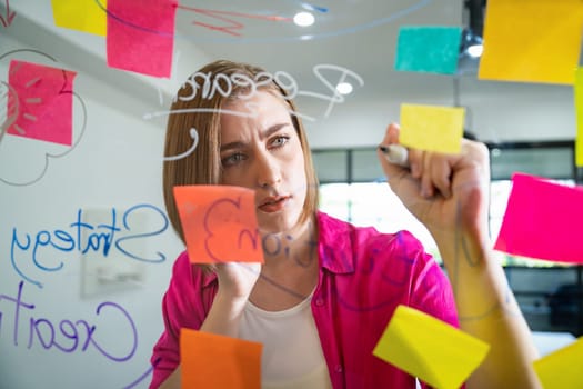 Closeup of young beautiful caucasian business leader presents marketing idea while writing marketing idea on glass board with mind map and colorful sticky notes. Portrait. Brainstorm. Immaculate.