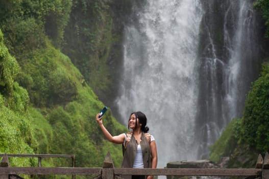 A cheerful woman captures a moment of her adventure with a selfie against the backdrop of a towering waterfall surrounded by vibrant green foliage.