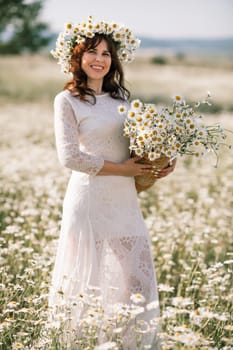 Happy woman in a field of daisies with a wreath of wildflowers on her head. woman in a white dress in a field of white flowers. Charming woman with a bouquet of daisies, tender summer photo.