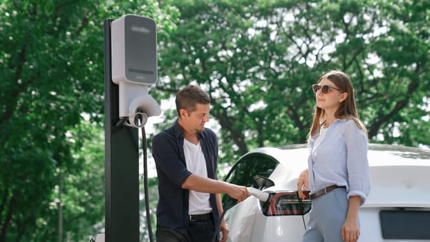 EV car recharging electricity for battery by lovey couple traveling during their road trip travel by eco friendly electric vehicle in national park and greenery forest on holiday. Exalt