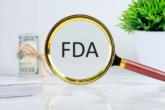 Word FDA written through a magnifying glass on a light background near a roll of money and a pot of green grass