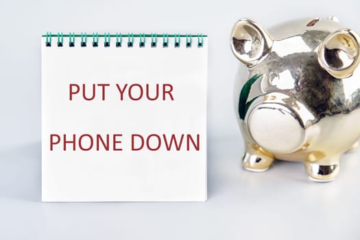 Put Your Phone Down text written on a notebook on a gray background next to a piggy bank