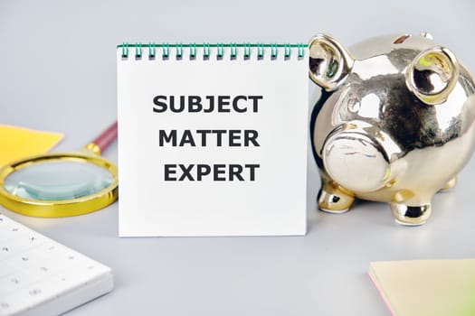 Text Subject Matter Expert written on a notebook on a gray background near a calculator, a piggy bank, and stickers. Small Business Concept