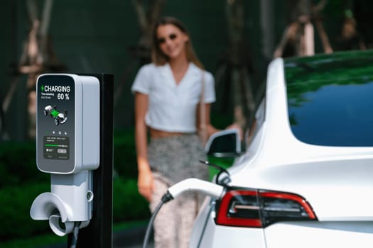 Young woman travel with EV electric car charging in green sustainable city outdoor garden in summer. Urban sustainability lifestyle by green clean rechargeable energy of electric BEV vehicle innards