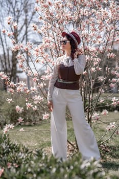 Magnolia park woman. Stylish woman in a hat stands near the magnolia bush in the park. Dressed in white corset pants and posing for the camera