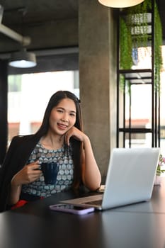 Portrait of millennial businesswoman holding cup of coffee and smiling at camera.