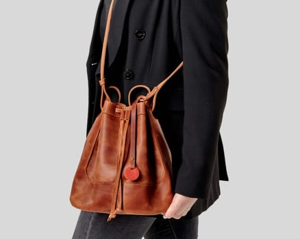 Close-up of stylish woman wearing casual black jacket and jeans holding chic brown leather drawstring bag over shoulder against neutral background