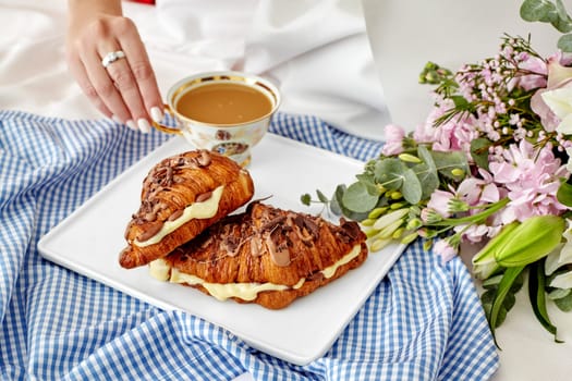Romantic intimate breakfast in bed scene with woman reaching for cup of coffee, fresh French croissants with delicate custard and chocolate, and lush flower bouquet on white sheets, cropped shot
