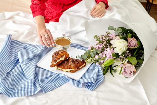 Elegant romantic morning setting with woman in red enjoying breakfast in bed with fresh French chocolate croissants filled with delicate custard and tender flower bouquet on white sheets, cropped shot