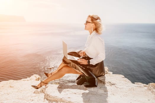 Successful business woman in yellow hat working on laptop by the sea. Pretty lady typing on computer at summer day outdoors. Freelance, travel and holidays concept.