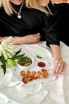 Sophisticated woman relaxing on white bedding with tray of delicious freshly baked sausage rolls, healthy vegetable salad, spicy dipping sauces, and fragrant white lilies. Romantic weekend concept