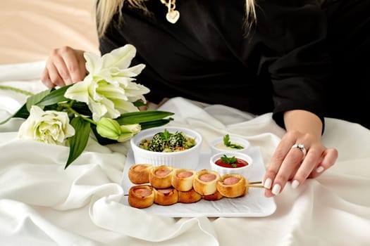 Woman in black outfit enjoying romantic gourmet snack of sausage rolls on skewers, fresh vegetable salad and piquant dipping sauces, lying on bed beside white lilies. Cropped image