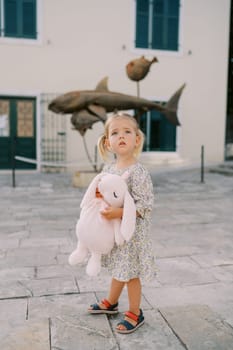Little girl stands hugging a pink toy rabbit in the courtyard of an old building and looks up. High quality photo