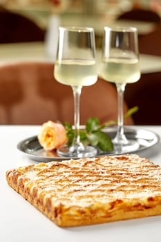 Romantic dining scene with two glasses of white wine and rose served on tray in blurred background and freshly baked golden lattice cheese tart, set for intimate gourmet experience