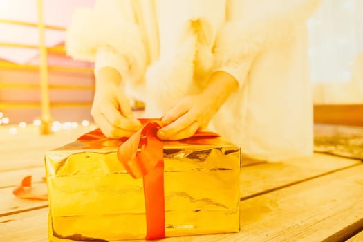 A woman in a white dress is holding a gold box with a red ribbon. She is wearing a crown on her head. The scene takes place in a room with a door and a window. The woman appears to be opening the gift box.