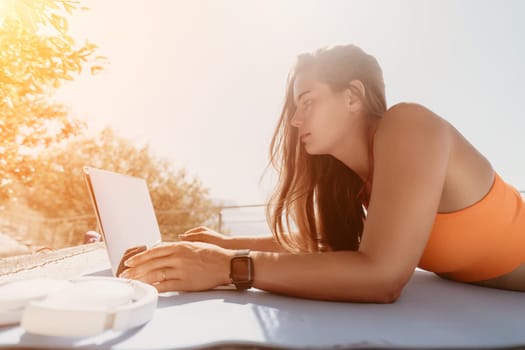 Digital nomad, Business woman working on laptop by the sea. Pretty lady typing on computer by the sea at sunset, makes a business transaction online from a distance. Freelance, remote work on vacation