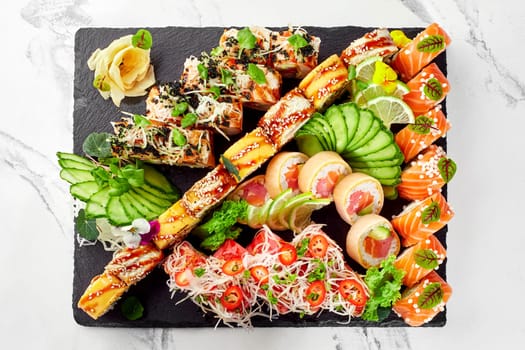 Top view of lavish assortment of salmon, eel and tuna sushi rolls displayed on slate board and garnished with fresh cucumber and lime slices, flowers, sliced spicy chili peppers and shredded daikon