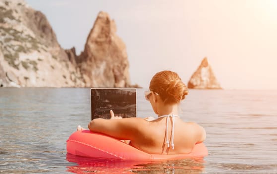 Woman freelancer works on laptop swimming in sea on pink inflatable ring. Pretty lady typing on computer while floating in the sea on inflatable donut at sunset. Freelance, remote work on vacation