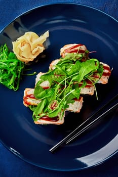 Top view of gourmet sushi roll with salmon seasoned with spicy mayo and unagi sauce topped with fresh greens on deep blue plate, accompanied by pickled ginger, seaweed salad and chopsticks