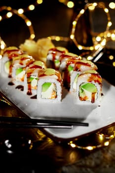 Enticing plate of sushi rolls filled with cream cheese, tobiko and avocado, topped with scallop, drizzled with sauces, served in festive setting with warm lights