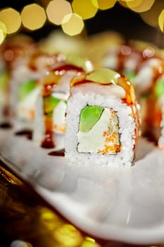 Closeup of sushi roll filled with cream cheese, tobiko, cucumber and avocado, topped with sea scallop and drizzled with sauces, served on plate in festive setting with warm lights