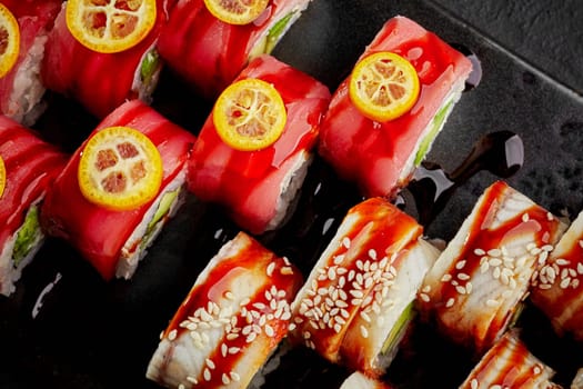 Top-down image showcasing vibrant sushi rolls topped with tuna and eel garnished with slices of kumquat and sesame seeds, seasoned with tangy unagi sauce on dark slate