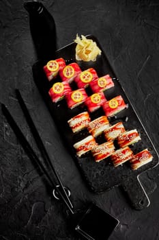 To view of set of rolls with eel and tuna dressed with unagi sauce, sesame and slices of kumquat on black serving board with pickled ginger and soy sauce. Traditional Japanese cuisine