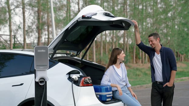 Couple recharge their EV car with electric battery charging station at rest stop during their road trip travel during autumnal season to national park. Relaxing holiday with eco friendly car. Exalt