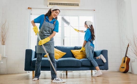 Two smiling women in uniform in messy living room working a team for house cleaning service. Using professional equipment they care for furniture sofa and hygiene. Housework is their routine.