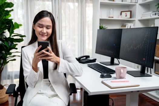 Smiling young Asian businesswoman with happy face looking on smartphone in profitable of exchange stock market graph at modern office. Concept of investing high profit in wealth security. Stratagem.